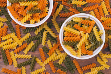 Three-colored uncooked pasta in the ceramic bowls on the wooden background with scattered pasta on it. Top view.