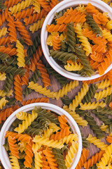 Three-colored uncooked pasta in the ceramic bowls on the wooden background with scattered pasta on...