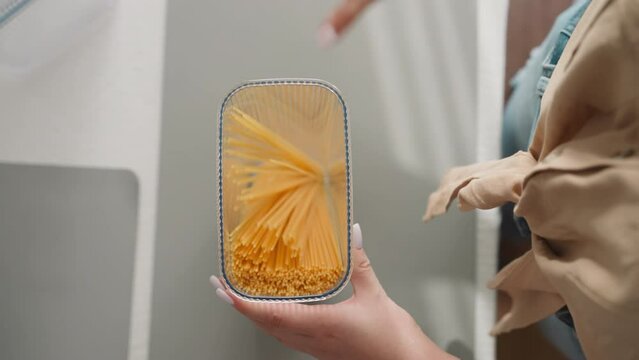 Woman puts spaghetti into transparent plastic container and closes lid shaking upper view. Young lady packs raw food into convenient box for storage