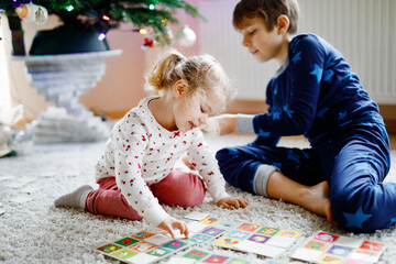 Two little chilren, cute toddler girl and school kid boy playing together card game by decorated Christmas tree. Happy healthy siblings, brother and sister having fun together. Family celebrating xmas