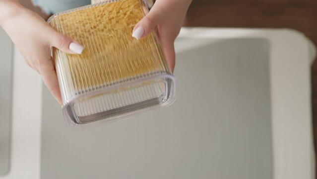 Female hand shakes closed container with spaghetti over kitchen table with serving mat slow motion. Lady packs raw food into convenient package for storage upper view