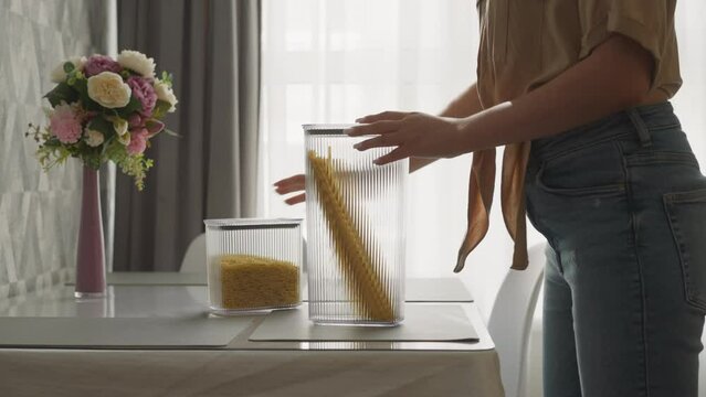 Housewife examines packaged raw spaghetti slow motion. Woman stacks containers on table with bouquet evaluating convenience of using plastic kitchen utensils