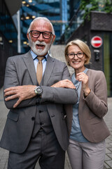 Portrait of senior man dressed in a suit and a mature blonde woman as business partners husband and wife friends or colleagues stand in front of modern building in day