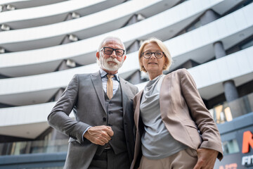 Fototapeta premium Portrait of senior man dressed in a suit and a mature blonde woman as business partners husband and wife friends or colleagues stand in front of modern building in day