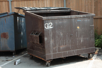 empty garbage dumpster with lid open and another dumpster behind it, surrounded by wood fence in...