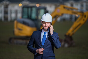 Construction owner near excavator. Confident construction owner in front of house. Architect, civil engineer. Man construction owner with a safety vest and hardhat at construction site.