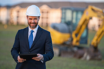 Construction manager in suit and helmet at a construction site. Construction manager worker or supervisor wearing hardhat in front of house. Supervisor construction manager near excavator. Renovation.