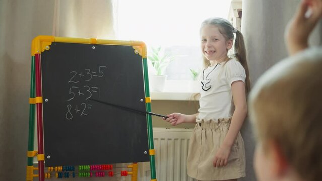 Smiling girl asks little brother to solve maths tasks standing by chalkboard at home. Elder sister teaches toddler boy mathematics lesson in children room