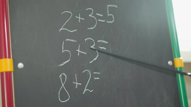 Teacher shows maths tasks written on chalkboard with pointer in classroom closeup. Simple mathematical problems for primary school students on blackboard
