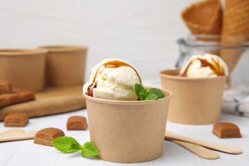 Scoops of ice cream with caramel sauce, mint leaves and candies on white tiled table, closeup