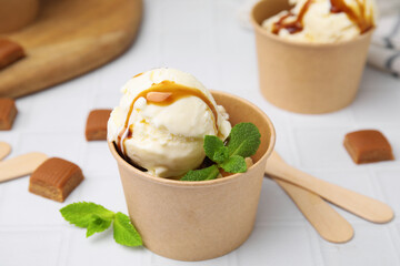 Scoops of ice cream with caramel sauce, mint leaves and candies on white tiled table, closeup