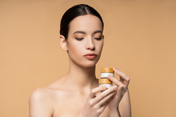 Brunette asian woman with naked shoulders holding jars with cosmetic cream isolated on beige