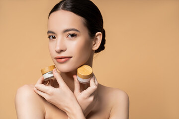 Young asian woman with naked shoulders holding jars with cosmetic cream and posing isolated on beige