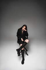 Trendy young asian woman in black jacket sitting on chair and posing on grey background