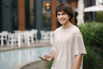 Teenage boy listen music in wireless headphones and hold his smartphone. Happy male. Background of cafe and park