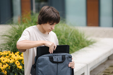 Boy open laptop bag and takes out his laptop. Boy sits on bench outdoors