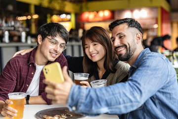 Multiracial friends toasting glasses of beer sitting in the brewery pub while taking a selfie with their smart phone