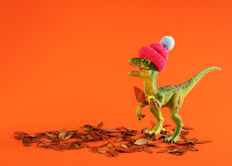 Cute green toy dinosaur wearing knitted hat and holding autumn leaf on orange background.