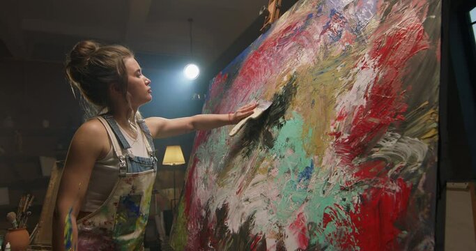 Millennial girl artist paints picture in loft studio using spatula to apply paint to canvas. Abstraction cinematic. Female creative artist concentrated and inspired while working on new masterpiece.