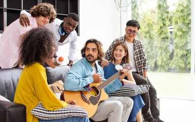 Group of diverse friends or people wearing casual clothes, relax smiling with happiness, party celebration, sitting in living room at cozy home, playing guitar and singing. Birthday, New Year Concept.