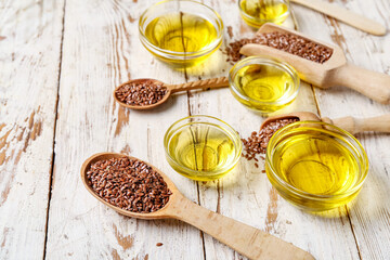 Glass bowls of flax oil and spoons with seeds on white wooden background