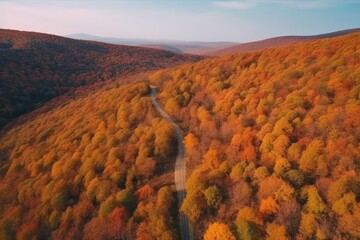 Top view of a winding road through a wooded mountain range in autumn with orange, yellow and red trees, a blue sky with clouds and dark blue mountains disappearing into the horizon.