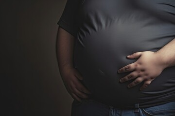 Belly of a fat man in a gray t-shirt and blue jeans, with his hand on his stomach, against a dark background.