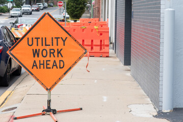 Bright orange and black, diamond shaped, utility work ahead sign on a city sidewalk. 