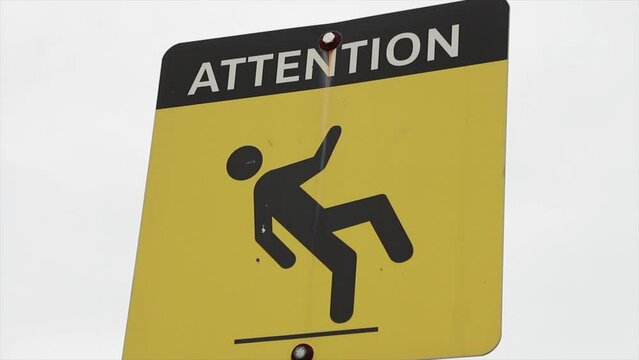 yellow square rectangle sign that says attention in capital letters with an illustrative picture of a person slipping and falling on a flat line surface, black and white print, on metal post