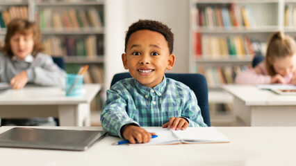 Portrait of funny black pupil sitting at desk in classroom at the elementary school and looking at camera