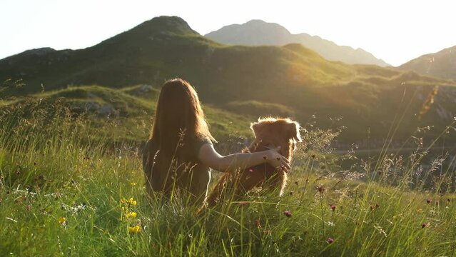 The girl strokes her dog Nova Scotia tolling retriever while sitting in the tall grass in the meadow. The owner and pet watch the sunset. National park Durmitor in Montenegro.