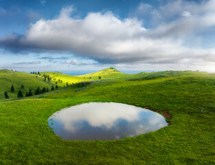 Obraz premium Aerial view of pond and green alpine meadows and hills at sunset in summer. Top drone view of mountain valley, green grass and sky with clouds reflected in water. Velika Planina, Slovenia. Landscape