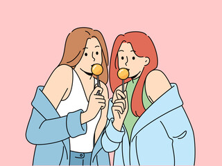 Two women with chupa chups in hands dressed in fashionable seductive clothes eat lollipops. Lovely girls in stylish jackets slung off shoulders to advertise new candies for teenagers