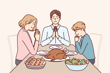 Family prayer before dinner, sitting at table with delicious food and thank god for well-being. Happy religious man and woman together with son say prayer, observing catholic ritual.