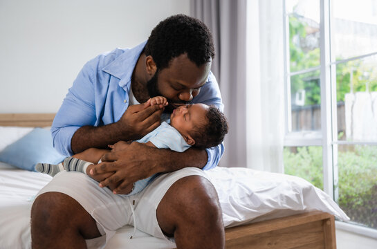 Happy African American father man sitting holding and kissing newborn son baby on a bed in the bedroom at home.