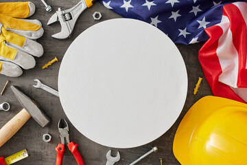 Paying homage to laborers on Labor Day. Top view flat lay of national flag, building instruments, helmet, work gloves on concrete grey background with blank circle for advert or text