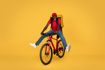 Glad young african american delivery man in uniform with big backpack and open mouth, has fun, riding bike