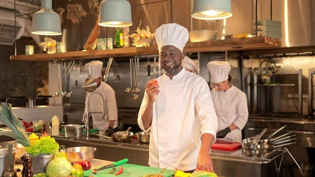 African American male chef standing and juggles tomato. Multicultural cook stands at background of restaurant's kitchen, looking at camera and smiling. Proffesional group of cooks working together.
