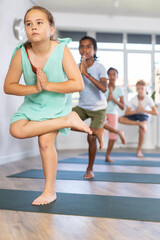 Children practicing yoga, standing in Tree Pose, asana Vriksasana, leg in half lotus position, hands in Namaste.