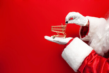 santa claus in suit and gloves holds empty shopping cart on red background, empty small cart stands...