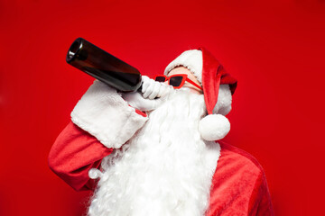 drunk santa claus in hat and festive glasses drinks wine from bottle on red background, man in santa costume