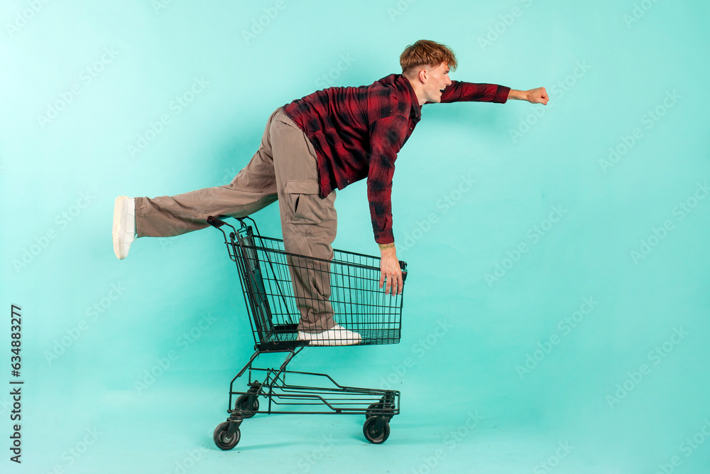 Wall mural young happy guy shopper sits and rides in shopping cart from supermarket on blue isolated background