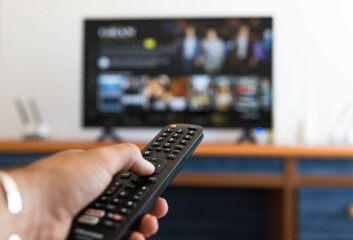 Hand holding remote control while searching for a serie on television - obrazy, fototapety, plakaty