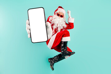 santa claus in sunglasses and suit jumps and flies and shows blank smartphone screen on blue...