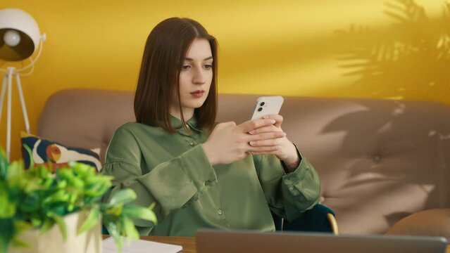 Portrait of serious woman using mobile phone in room. Focused girl typing text on smartphone. Female student resting after lessons. Young lady sitting at the table with laptop computer, 4k footage