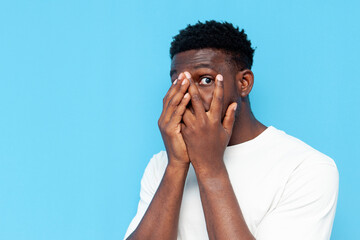 frightened african american man in white t-shirt covers his face with his hands and peeps on blue isolated background