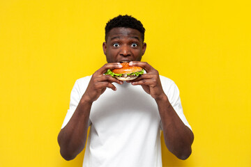 shocked african american man in white t-shirt holding big burger and surprised, amazed guy eating...