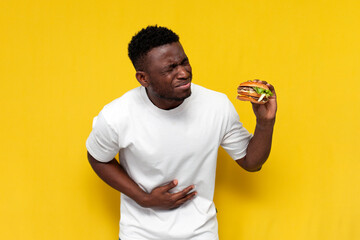 unhappy african american man in white t-shirt eats spoiled fast food and holding on to his stomach