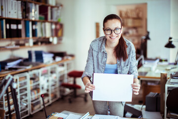 Young woman holding a blank piece of paper in a startup company office with copy space