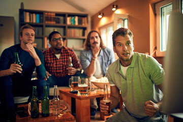 Young group of male friends watching a sports game in the living room on the tv and cheering for their team